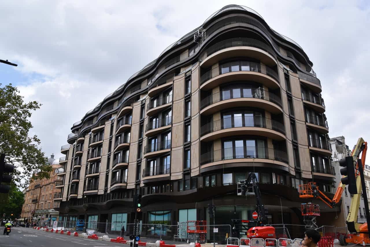 Curved multi-storey residential building with a dark brick and stone facade and rows of balconies, with construction equipment at the edge.