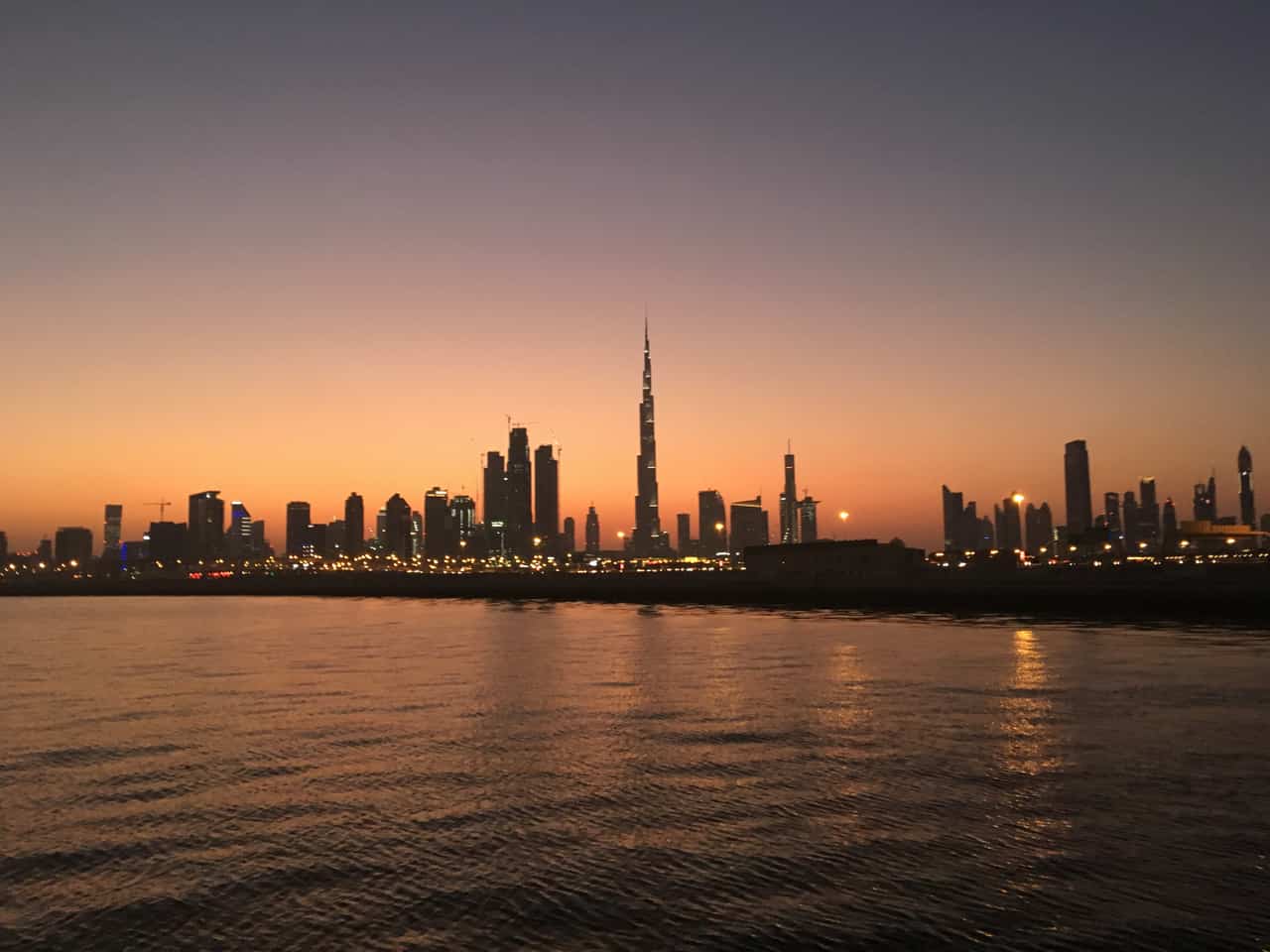Very tall skyscraper silhouetted against an orange dusk sky above a city skyline reflected in calm water.