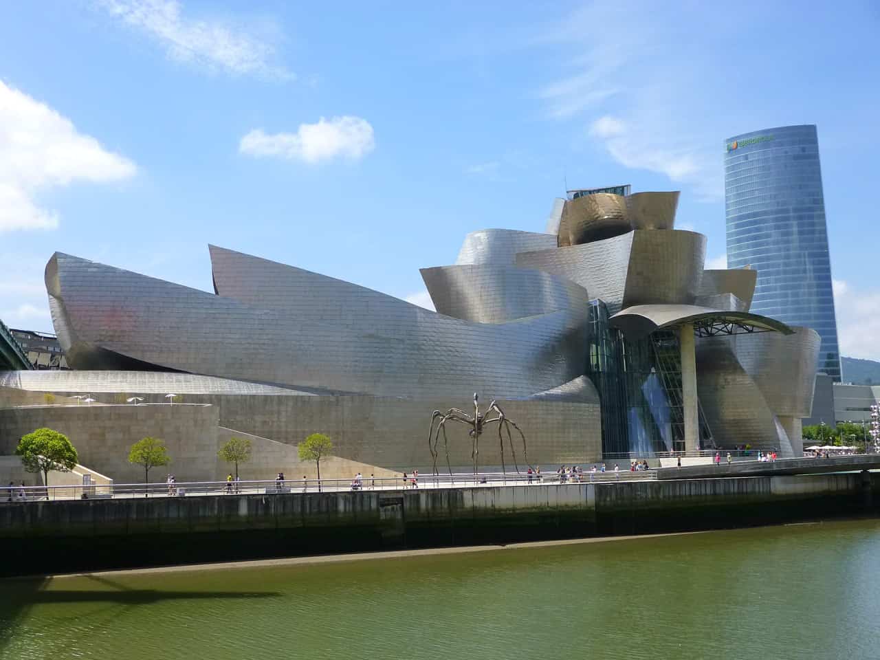 Curving titanium-clad museum building beside a river, with a tall glass tower behind it and blue sky overhead.
