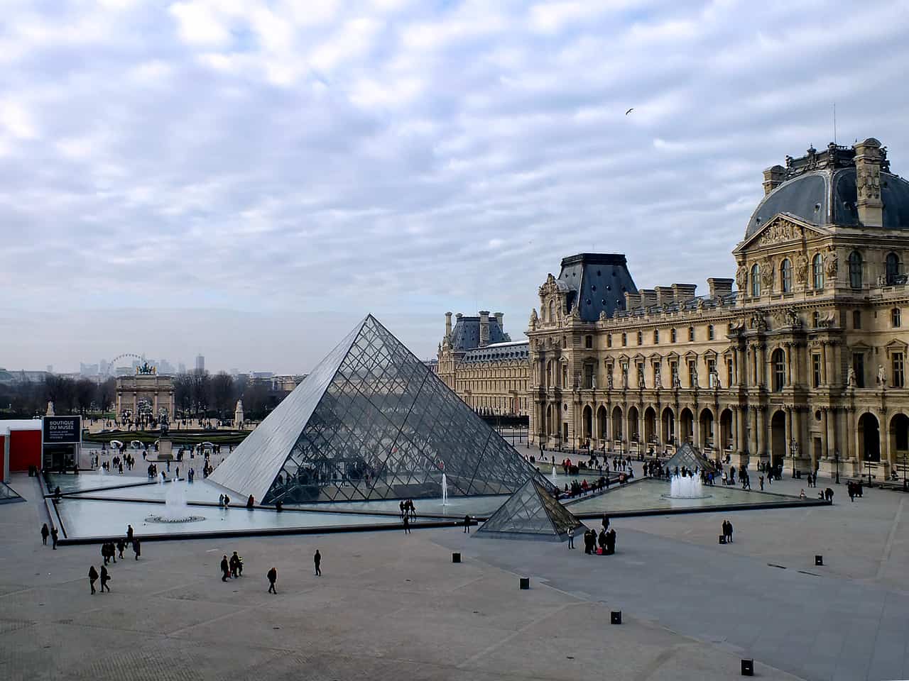 Glass pyramid in the courtyard of a large classical palace under a cloudy sky.