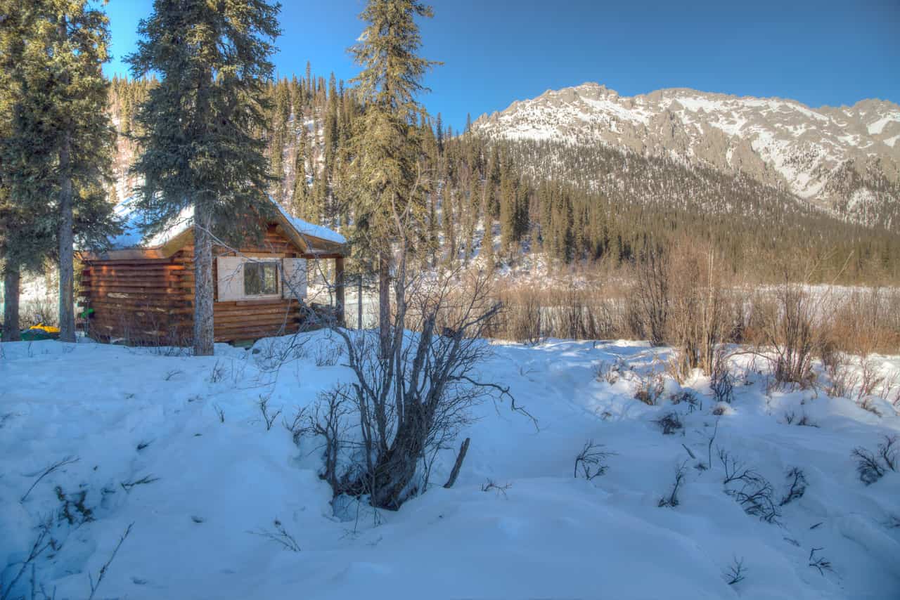 Small log cabin among spruce trees in deep snow with snow-covered mountains rising in the background.