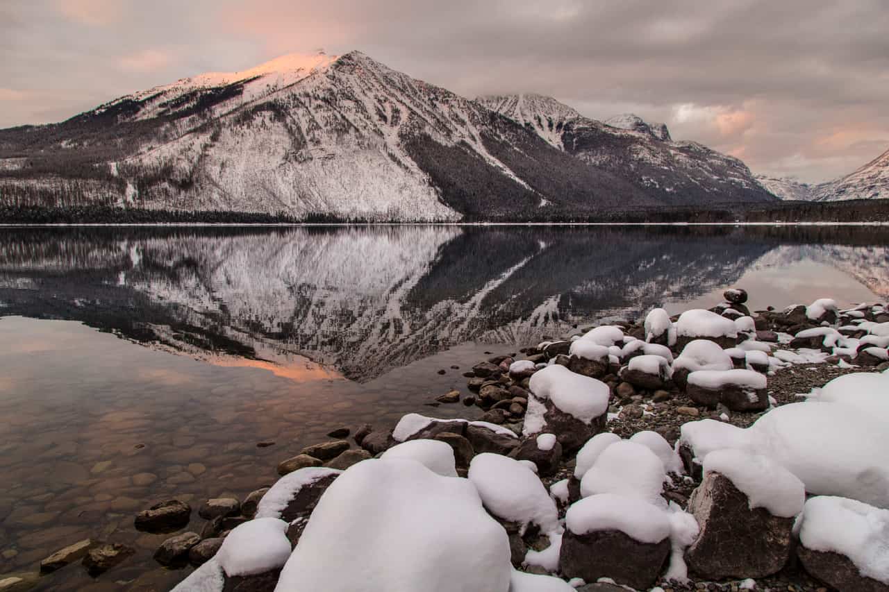 Snow-dusted mountain reflected in a still lake at sunset, with snowy rocks along the shoreline in the foreground.