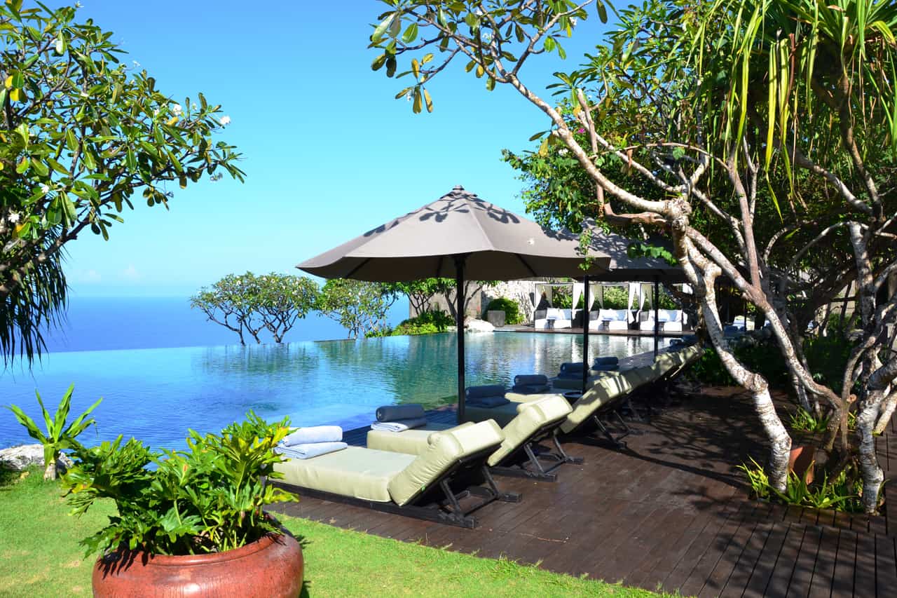 Resort infinity pool edge with cushioned sun loungers, a large parasol and tropical trees, with the sea visible beyond.