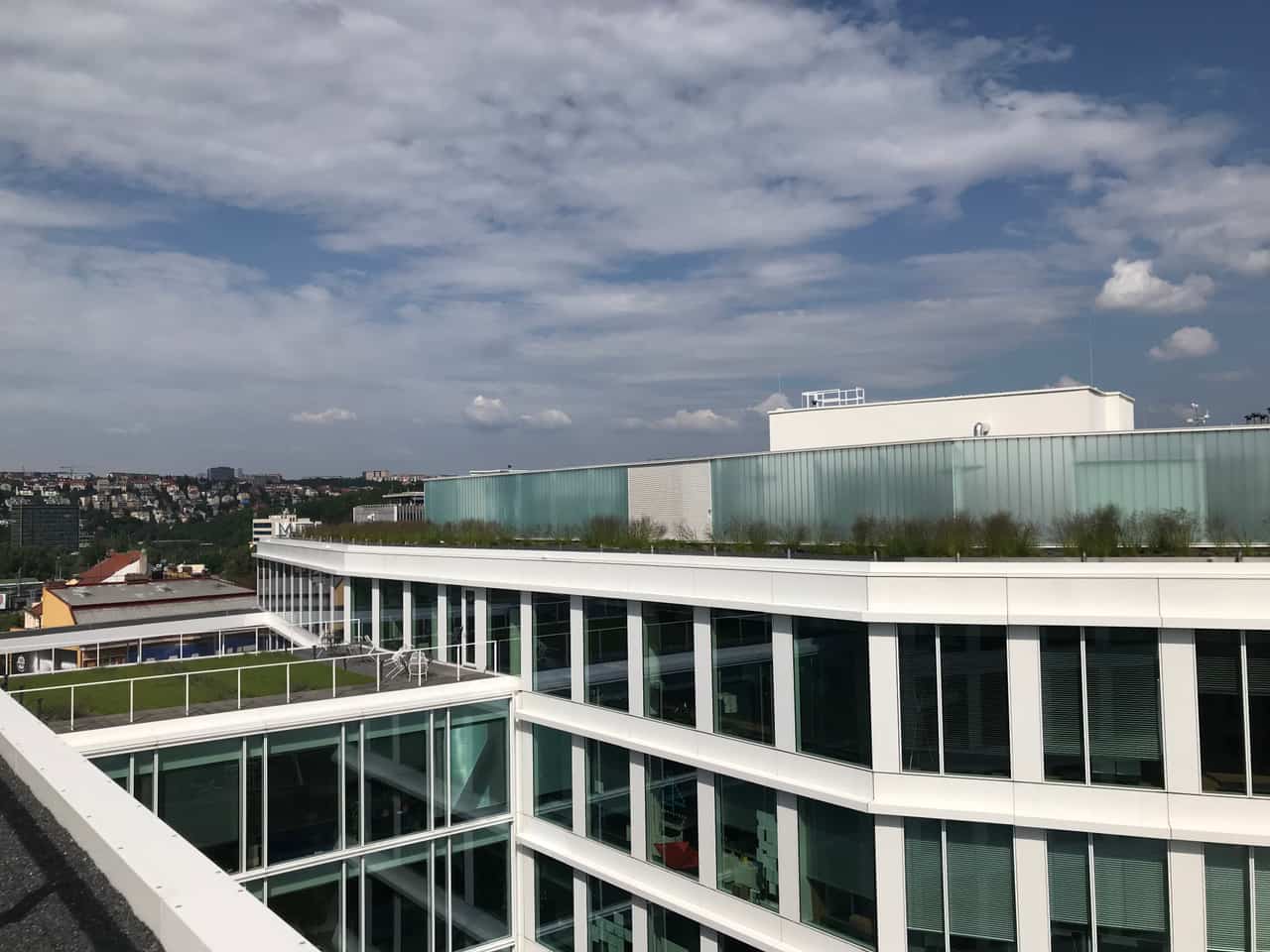 White modern office building with planted green terraces on its roof edge against a cloudy sky.