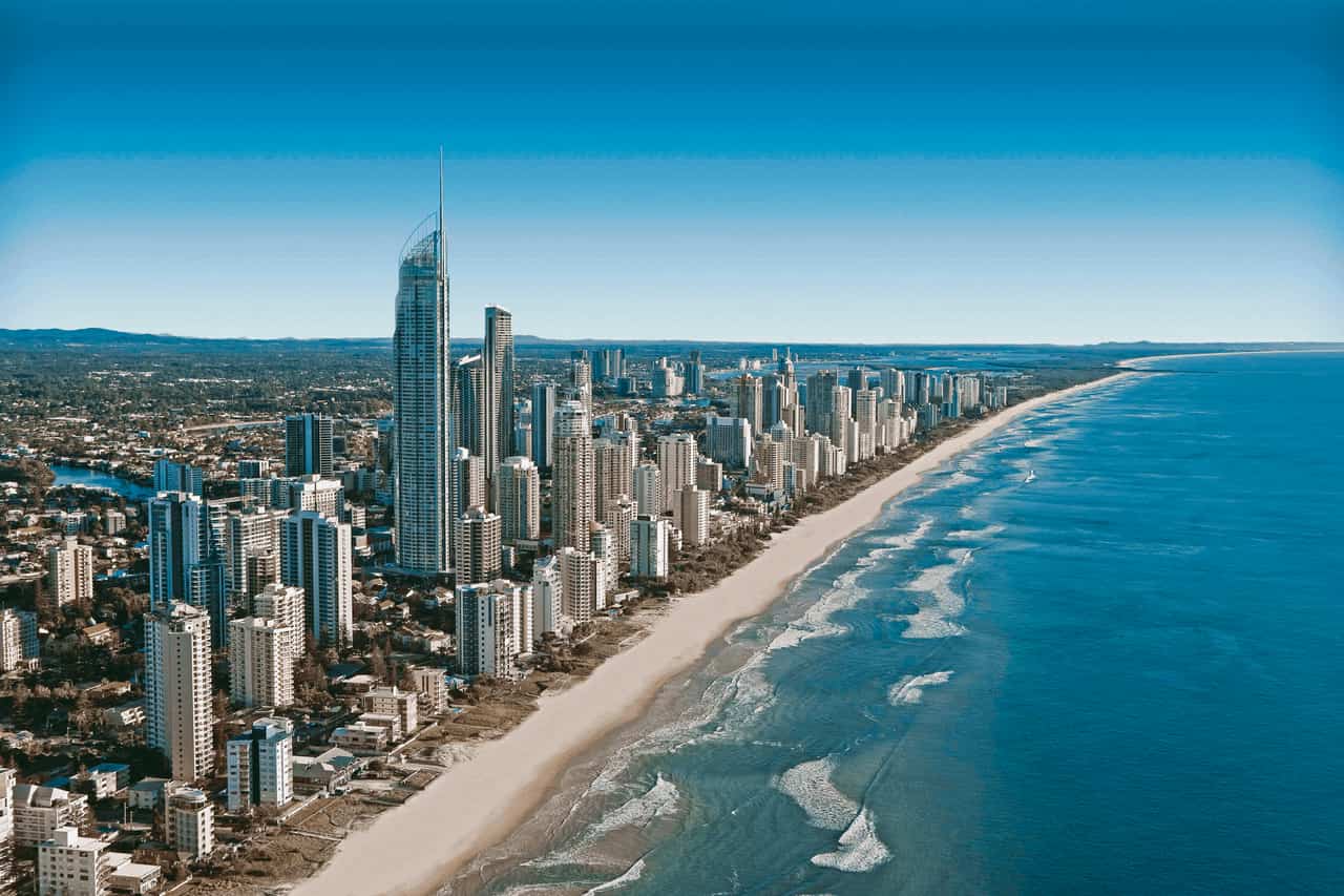 Aerial view along a long sandy beach with breaking waves and a dense row of high-rise apartment towers under a blue sky.
