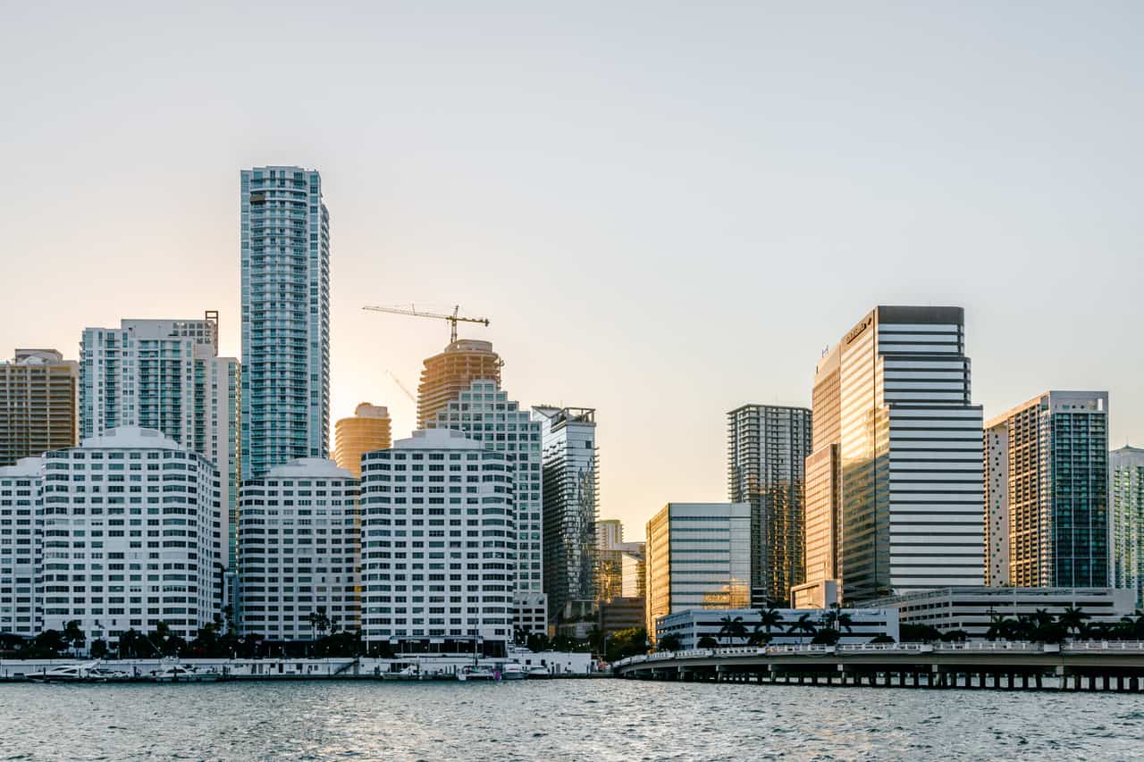 Downtown waterfront skyline of white and glass high-rise buildings with a cluster of cranes, seen across the water in daylight.