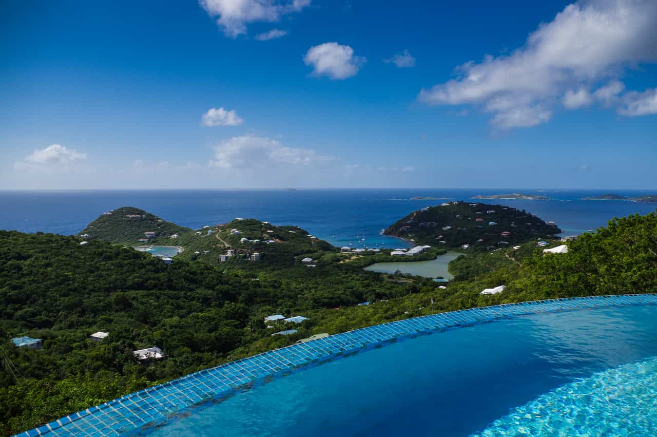 Blue infinity pool in the foreground overlooking green forested hills, a wide bay and the open sea under a partly cloudy sky.