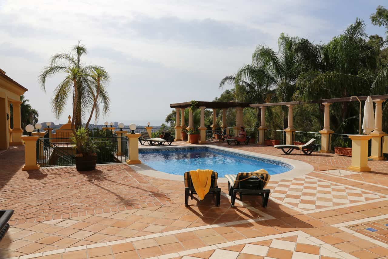 Terracotta paved pool terrace with a kidney-shaped swimming pool, sun loungers, palm trees and a pergola, with a hazy coastal town visible in the distance.